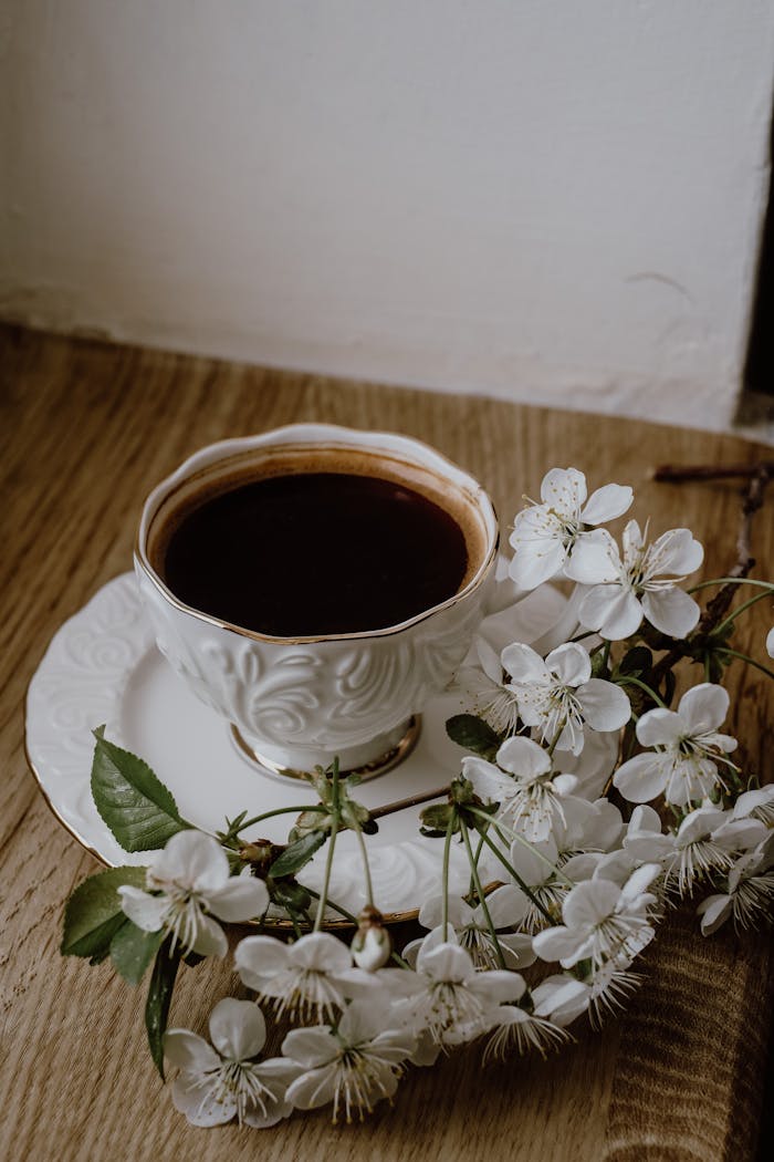 services-04 Ornate coffee cup and saucer surrounded by white cherry blossoms on a wooden table.